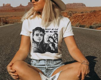 May include: Cream-colored t-shirt featuring a black and white image of a group of people, with the text "SLEEP ALL DAY PARTY ALL NIGHT". The shirt is paired with denim shorts and white cowboy boots.
