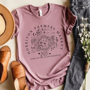 Pode incluir: Uma camiseta malva com uma ilustra&ccedil;&atilde;o em preto e branco de uma toca de hobbit com o texto "Hobbiton Farmers Market Home of the Second Breakfast".
