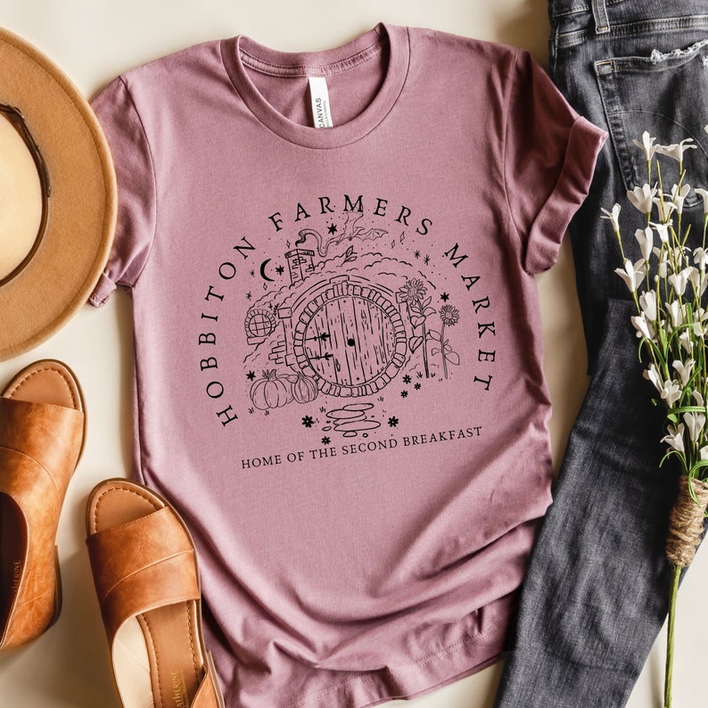 Pode incluir: Uma camiseta malva com uma ilustra&ccedil;&atilde;o em preto e branco de uma toca de hobbit com o texto "Hobbiton Farmers Market Home of the Second Breakfast".