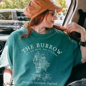 May include: Teal blue t-shirt with a white graphic of a house with the text "THE BURROW it's not much, but it's home Ottery St Catchpole, England"