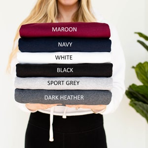 May include: A stack of folded sweatshirts in various colors, including maroon, navy, white, black, sport grey, and dark heather. Each sweatshirt is labeled with its color name. The sweatshirts are held by a person.