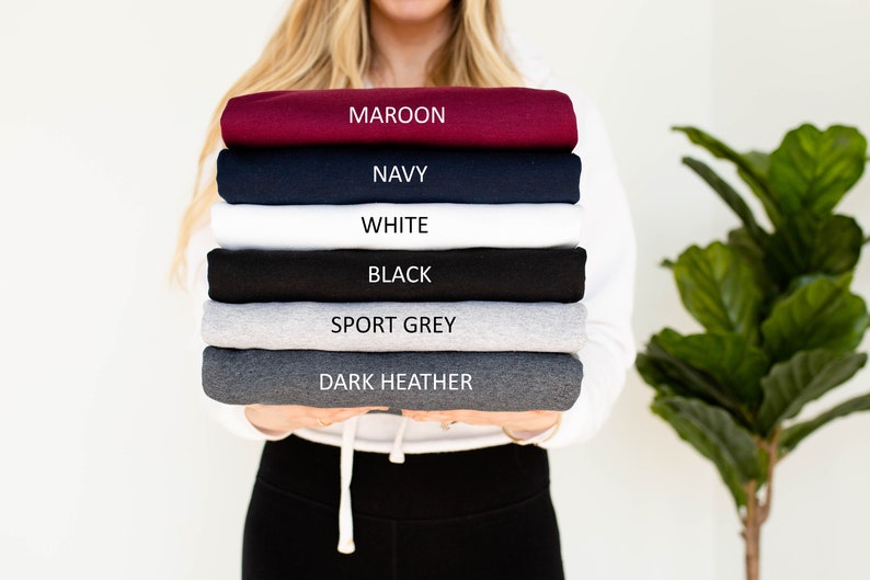 May include: A stack of folded sweatshirts in various colors, including maroon, navy, white, black, sport grey, and dark heather. Each sweatshirt is labeled with its color name. The sweatshirts are held by a person.