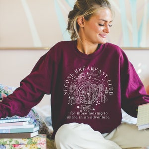 May include: Burgundy sweatshirt with a white graphic that reads "SECOND BREAKFAST CLUB" around a drawing of a hobbit door. The text "for those looking to share in an adventure" is below the graphic. The sweatshirt is being worn by a person.