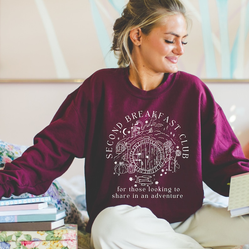 May include: Burgundy sweatshirt with a white graphic that reads "SECOND BREAKFAST CLUB" around a drawing of a hobbit door. The text "for those looking to share in an adventure" is below the graphic. The sweatshirt is being worn by a person.