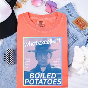 Op de afbeelding: Een oranje Comfort Colors sweatshirt met een afbeelding van een man met een hoed en de woorden "what excellent" en "BOILED POTATOES". Het sweatshirt ligt op een witte ondergrond met accessoires zoals een zonnebril en een scrunchie.