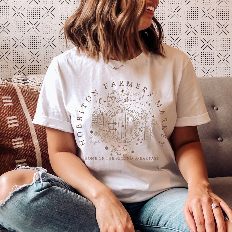 Pode incluir: Camiseta branca com um gr&aacute;fico marrom de um buraco de hobbit com o texto "Hobbiton Farmers Market" e "Home of the Second Breakfast".