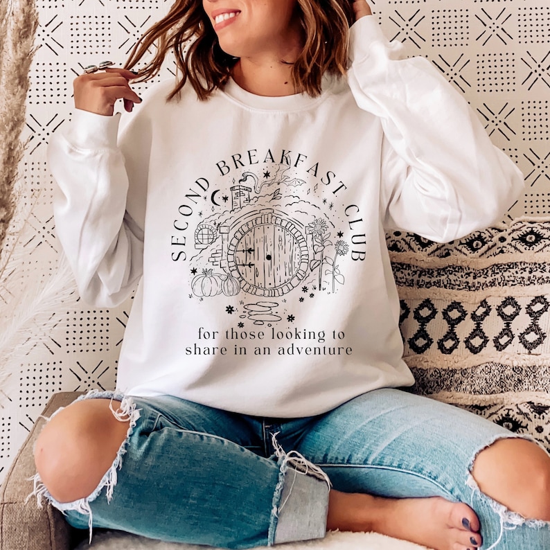 May include: White sweatshirt with a black graphic of a round door, pumpkins, and flowers, with the text "SECOND BREAKFAST CLUB" and "for those looking to share in an adventure". The sweatshirt has long sleeves.