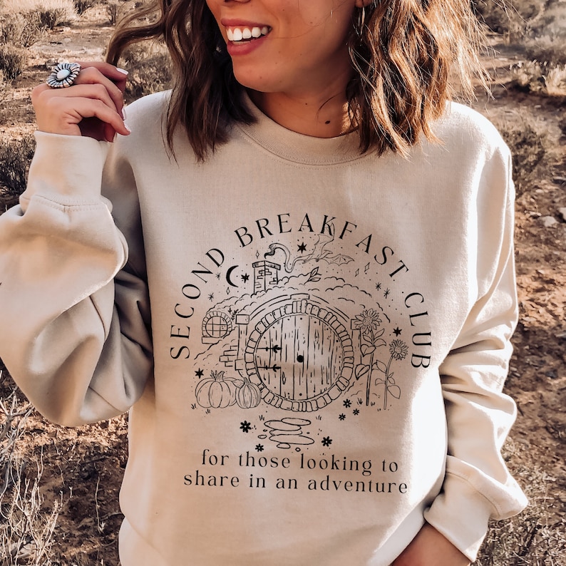 May include: Beige sweatshirt with the text "SECOND BREAKFAST CLUB" surrounding a graphic of a round door, pumpkins, and flowers. Below the graphic is the text "for those looking to share in an adventure."