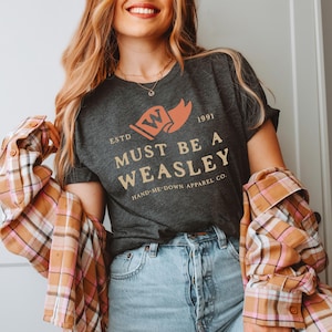 May include: Dark gray t-shirt with the text "MUST BE A WEASLEY" in a vintage style font. The shirt features a graphic of a banner with the letter "W" and the year "1991". The shirt is part of the "HAND-ME-DOWN APPAREL CO." collection.