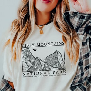 Op de afbeelding: Wit T-shirt met een zwart grafisch ontwerp van bergen en een adelaar. De tekst "Misty Mountains National Park" en "you'll have a tale or two to tell when you come back" staat onder het ontwerp gedrukt.