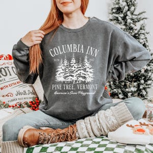 May include: A grey sweatshirt featuring the text "COLUMBIA INN" above a pine tree graphic, with "PINE TREE, VERMONT" and "America's Snow Playground" below. The sweatshirt is paired with jeans and boots.