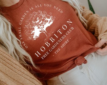 May include: A terracotta-coloured t-shirt with the text "ONE SMALL GARDEN IS ALL YOU NEED" and "HOBBITON FREE GARDENERS CLUB - THE SHIRE" in white. The shirt features a white tree graphic. The shirt is knotted at the waist.