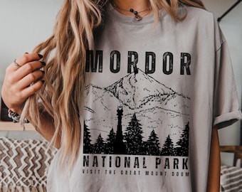 May include: Gray t-shirt with a black graphic of a mountain range, trees, and the text "MORDOR NATIONAL PARK VISIT THE GREAT MOUNT DOOM". The shirt has short sleeves and a relaxed fit.