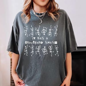 Puede incluir: Camiseta gris oscuro con el texto "It Was a Graveyard Smash" e ilustraciones de esqueletos bailando en blanco. La camiseta tiene cuello redondo y mangas cortas. Una prenda informal con temática de Halloween.