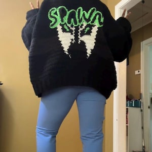 Puede incluir: Un cárdigan de crochet negro con un diseño de crochet verde y blanco en la espalda que dice "Spawn". La persona que lleva el cárdigan también lleva pantalones vaqueros azules.