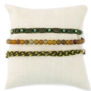 May include: Three green and brown bracelets on a white fabric background. The top bracelet is made of green string with green beads. The middle bracelet is made of brown beads. The bottom bracelet is made of green and brown string.
