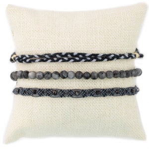 May include: Three bracelets stacked on a white pillow. The top bracelet is a black and white braided cord. The middle bracelet is made of gray gemstone beads. The bottom bracelet is a black braided cord with small silver beads.
