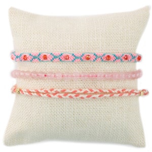 May include: Three pink and blue bracelets on a white pillow. The top bracelet is a woven design with pink, blue, and red threads. The middle bracelet is made of pink beads. The bottom bracelet is a woven design with pink and red threads.