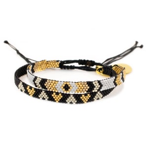 May include: Two beaded bracelets with black, white, and gold geometric patterns. The bracelets are tied together with a black cord and a gold charm.