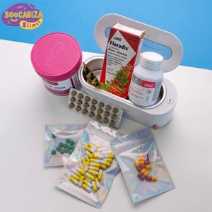 K&ouml;nnte beinhalten: Ein wei&szlig;er Pillenorganizer mit einem durchsichtigen Deckel, der verschiedene Pillen und Kapseln enth&auml;lt. Der Organizer ist ge&ouml;ffnet und hat einen kleinen Knopf an der Seite. Es gibt auch mehrere kleine Plastikt&uuml;ten, die mit Pillen und Kapseln gef&uuml;llt sind.