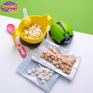 K&ouml;nnte beinhalten: Ein Set aus drei durchsichtigen Plastikt&uuml;ten, gef&uuml;llt mit kleinen, runden, wei&szlig;en und beigefarbenen Snacks. Die Beutel haben einen Rei&szlig;verschluss. Eine gelbe Sch&uuml;ssel mit Saugnapfbasis und einem Strohhalm ist mit den gleichen Snacks gef&uuml;llt. Ein rosa L&ouml;ffel und ein gr&uuml;nes Marienk&auml;fer-Spielzeug sind ebenfalls auf dem Bild zu sehen.