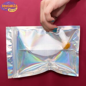 K&ouml;nnte beinhalten: Eine durchsichtige, regenbogenfarbene Plastikt&uuml;te mit Rei&szlig;verschluss. Eine Hand h&auml;lt die Oberseite der T&uuml;te offen. In der T&uuml;te befindet sich eine kleine, runde, orangefarbene Schleimb&auml;lle.