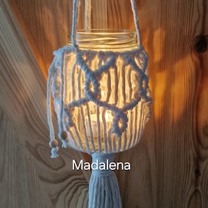 Könnte beinhalten: Eine handgefertigte Makramee-Laterne mit einem Glas im Inneren, die an einer weißen Schnur hängt. Die Laterne hat ein weißes Makramee-Design und eine weiße Quaste. Das Wort "Madalena" ist auf dem Glas aufgedruckt.