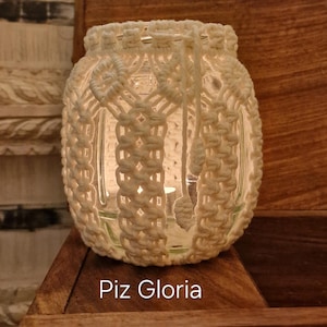 May include: A white macramé jar candle holder with a glass insert, on a wooden chest. The jar features an intricate woven geometric pattern. The text "Piz Gloria" is visible on the chest.