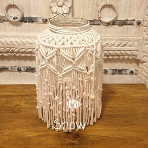 May include: A white macrame lampshade with wooden beads hangs from a glass jar. The lampshade is made with a knotting technique and has a fringe at the bottom. The jar is sitting on a wooden surface.