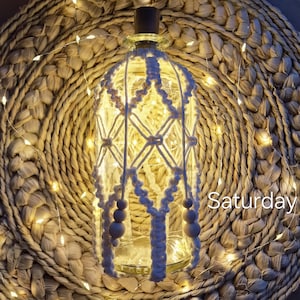 May include: A clear glass bottle adorned with white macrame and illuminated by warm white fairy lights. The bottle rests on a woven, circular placemat. The word "Saturday" is visible on the right side of the image.