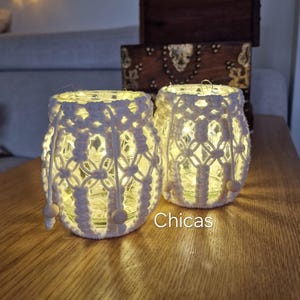 Puede incluir: Dos portavelas de cristal iluminados con fundas de macramé blanco. Los portavelas tienen un patrón decorativo y cuentas de madera. La palabra "Chicas" es visible en la mesa. Los portavelas están sobre una mesa de madera.