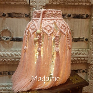 Puede incluir: Una pantalla de lámpara de macramé rosa con cuentas de madera y una cadena de luces en su interior. La pantalla está colgada de una estantería de madera con intrincados tallados.