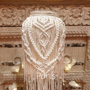 Op de afbeelding: Een witte macramé lampenkap met houten kralen die eronder hangen. De lampenkap heeft de vorm van een hart en heeft een geometrisch patroon. Het woord "Paris" staat op de lampenkap.