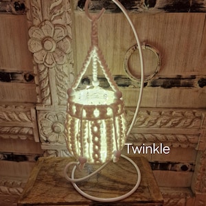 May include: A white macrame lantern, illuminated with a warm light, hanging from a curved metal stand. The lantern sits on a wooden surface, with the word "Twinkle" visible. The background features carved wooden panels.