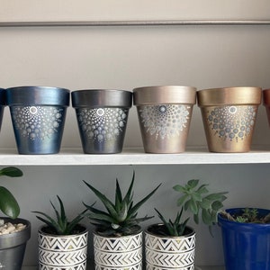 May include: Six painted terracotta pots with white geometric designs. The pots are arranged in a row on a shelf. The pots are in various colors, including blue, gray, brown, and copper.