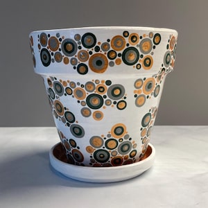 May include: White terracotta planter with a geometric pattern of gold, gray, and green circles. The planter has a matching saucer.
