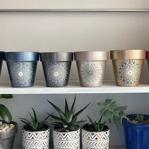 May include: Six terracotta flower pots with white mandala designs painted on them. The pots are arranged in a row on a white shelf. The pots are painted in different colors, including blue, gray, pink, gold, and copper.