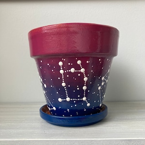 May include: A terracotta planter with a blue and pink ombre glaze and white painted constellations. The planter has a matching saucer.
