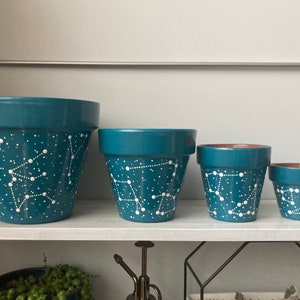 May include: Four teal blue terracotta pots with white painted constellations. The pots are arranged in a row from largest to smallest.