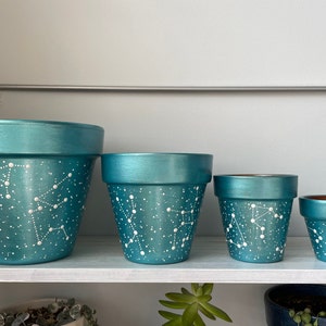 May include: Four teal green terracotta pots with white painted constellations and white dots. The pots are arranged in a row from largest to smallest on a white shelf.