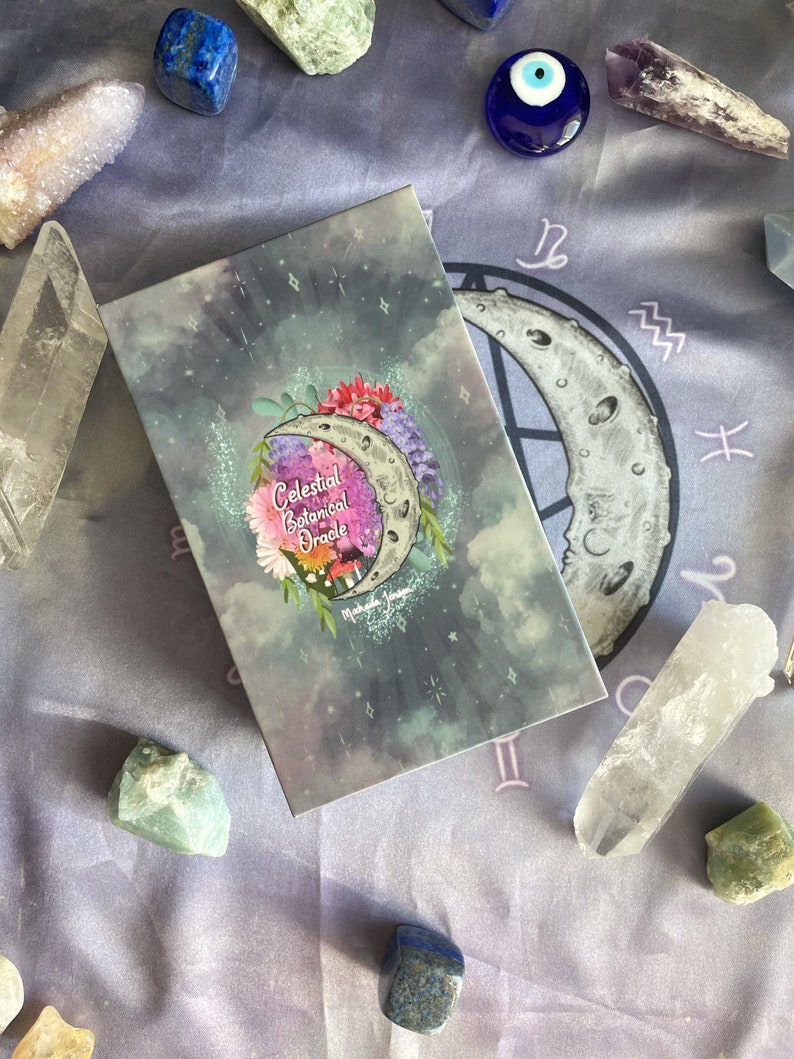 Puede incluir: Una caja gris con un dise&ntilde;o de or&aacute;culo bot&aacute;nico celestial. La caja est&aacute; rodeada de cristales y un amuleto azul de ojo turco. La caja tiene una luna creciente con flores y estrellas en ella. El texto en la caja dice "Celestial Botanical Oracle".