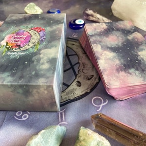 Puede incluir: Una baraja de cartas de Oracle Bot&aacute;nico Celestial con un dise&ntilde;o de cielo estrellado morado, rosa y azul. Las cartas est&aacute;n apiladas sobre un pa&ntilde;o morado con un s&iacute;mbolo de luna y zod&iacute;aco. La caja de las cartas tiene un dise&ntilde;o floral con una luna creciente y las palabras "Celestial Botanical Oracle" impresas en la parte delantera.