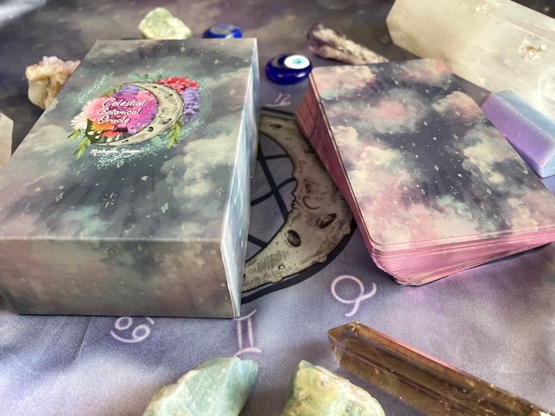 Puede incluir: Una baraja de cartas de Oracle Bot&aacute;nico Celestial con un dise&ntilde;o de cielo estrellado morado, rosa y azul. Las cartas est&aacute;n apiladas sobre un pa&ntilde;o morado con un s&iacute;mbolo de luna y zod&iacute;aco. La caja de las cartas tiene un dise&ntilde;o floral con una luna creciente y las palabras "Celestial Botanical Oracle" impresas en la parte delantera.