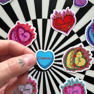 May include: A collection of heart-shaped stickers with various designs and text. One sticker is held by a hand, featuring a blue heart with the name "Cassian." Other stickers include zodiac signs and the word "Fireheart."