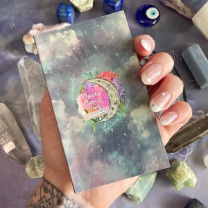 Puede incluir: Un peque&ntilde;o libro de gu&iacute;a azul y morado con un tema celestial y bot&aacute;nico. La portada presenta una luna creciente con flores y estrellas. El texto en la portada dice "Celestial Botanical Oracle Guidebook".