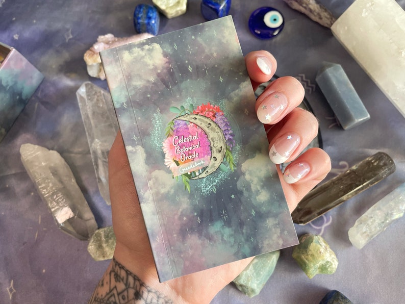 Puede incluir: Un peque&ntilde;o libro de gu&iacute;a azul y morado con un tema celestial y bot&aacute;nico. La portada presenta una luna creciente con flores y estrellas. El texto en la portada dice "Celestial Botanical Oracle Guidebook".