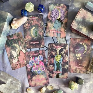 Puede incluir: Un juego de cartas de tarot con un tema celestial. Las cartas presentan una luna creciente, estrellas y dise&ntilde;os florales. Las cartas est&aacute;n dispuestas sobre un fondo de tela morado y gris con cristales y otros art&iacute;culos decorativos.