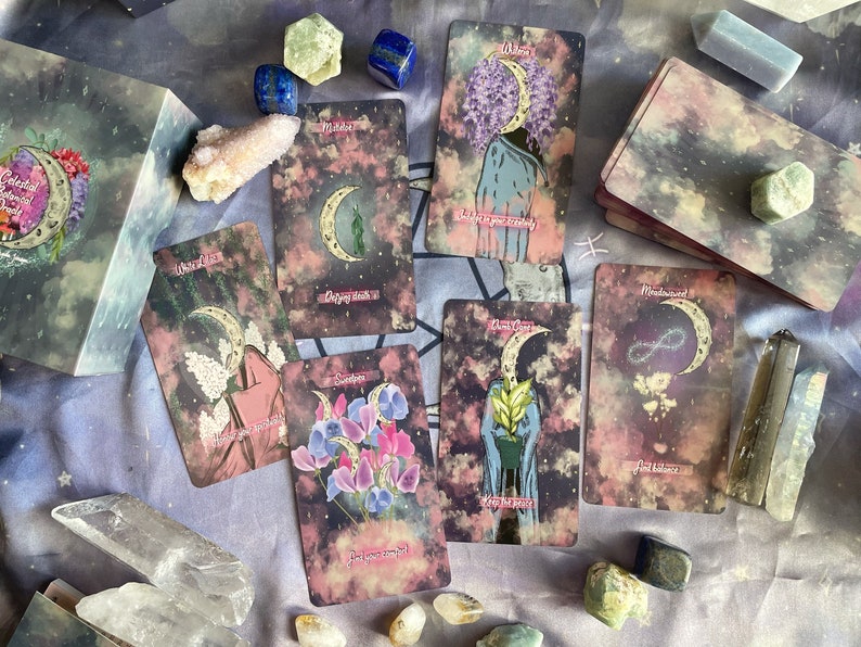 Puede incluir: Un juego de cartas de tarot con un tema celestial. Las cartas presentan una luna creciente, estrellas y dise&ntilde;os florales. Las cartas est&aacute;n dispuestas sobre un fondo de tela morado y gris con cristales y otros art&iacute;culos decorativos.