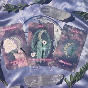Puede incluir: Tres cartas de tarot con un tema celestial. Las cartas est&aacute;n dispuestas sobre una tela de sat&eacute;n morado con cristales y vegetaci&oacute;n. Las cartas se titulan "Rom Chamomile", "Okra" y "Mulder", y presentan ilustraciones de una luna creciente, flores y una mano.