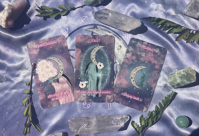 Puede incluir: Tres cartas de tarot con un tema celestial. Las cartas est&aacute;n dispuestas sobre una tela de sat&eacute;n morado con cristales y vegetaci&oacute;n. Las cartas se titulan "Rom Chamomile", "Okra" y "Mulder", y presentan ilustraciones de una luna creciente, flores y una mano.